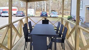 Un espacio de estar al aire libre techado, con mesa y sillas, un muelle de madera y vistas a coches aparcados y un edificio al fondo.