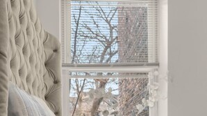Una cama con cabecera capitonada y manta a rayas, un jarrón blanco con flores en un alféizar, y vista a un árbol y un edificio.