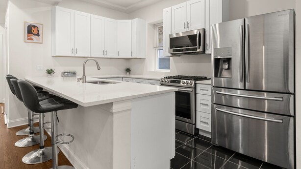 Una cocina moderna con armarios blancos, electrodomésticos de acero inoxidable, una isla central con taburetes y un microondas sobre la estufa.