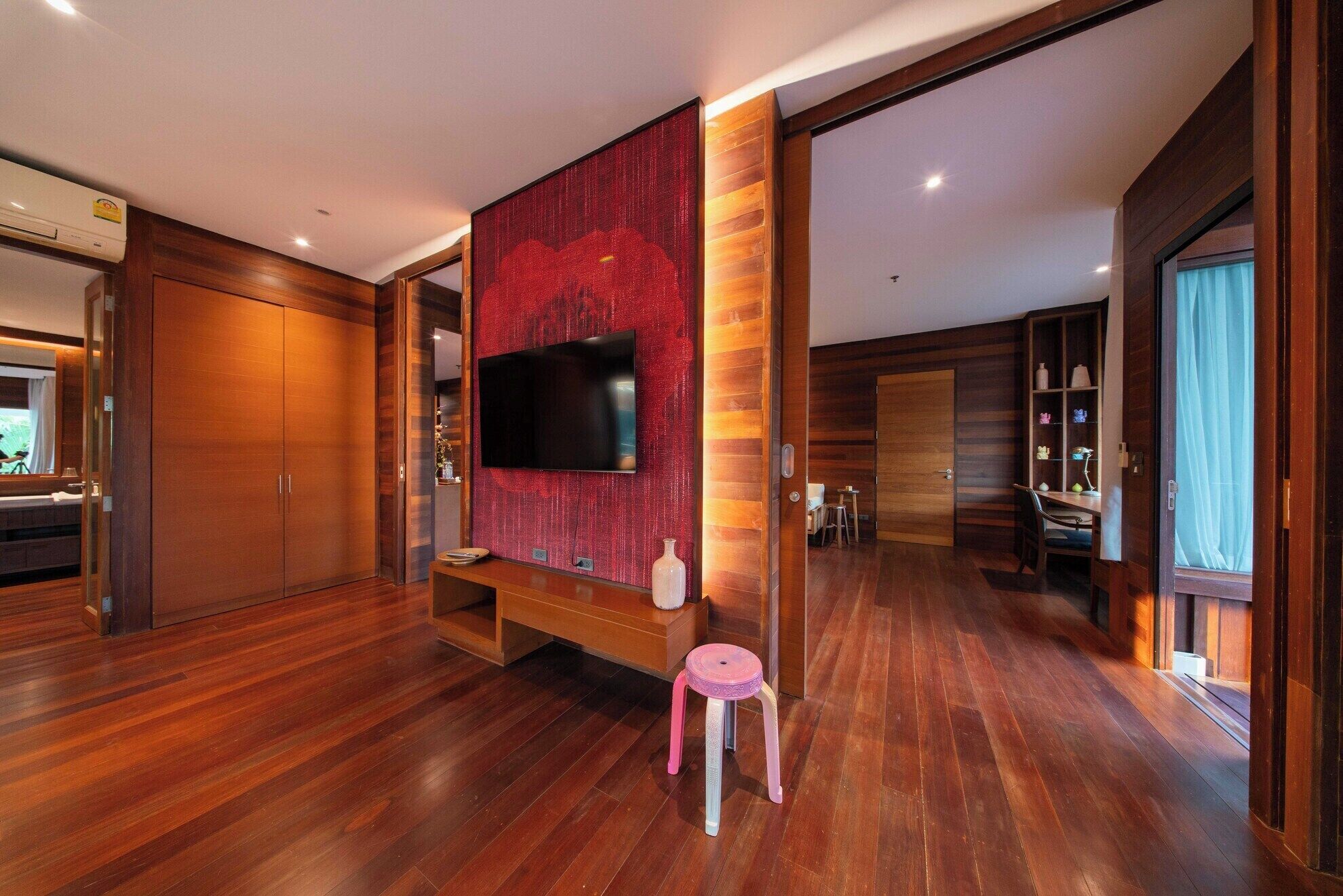 A spacious living room with a large flat-screen TV mounted on a red textured wall, wooden flooring, a small pink stool, and a dining area visible through an open door.