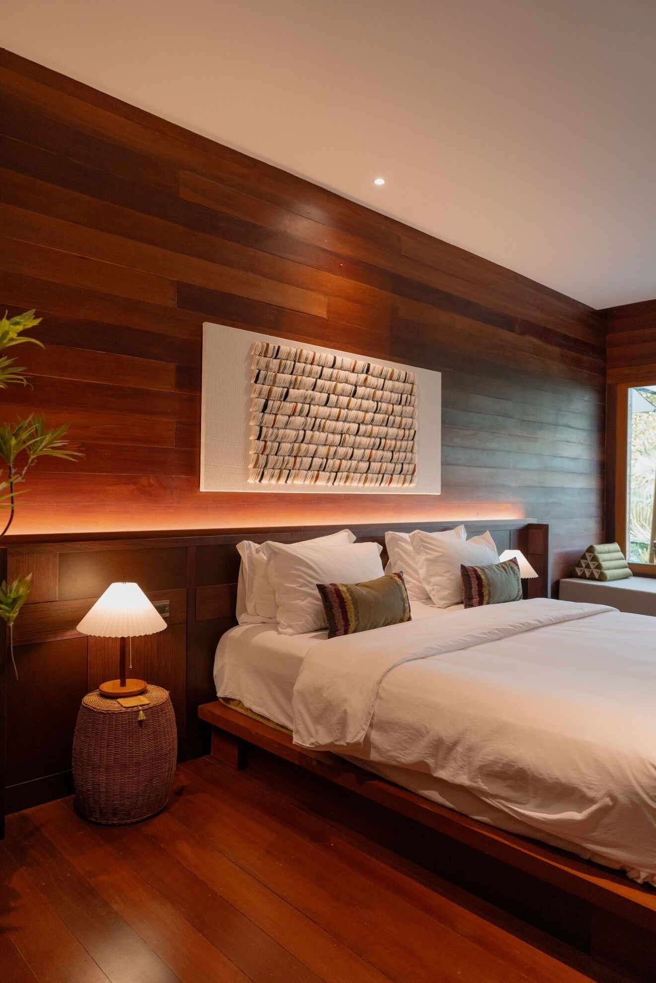 A bedroom with a large bed, wooden panel walls, a woven side table, and a wall-mounted artwork.