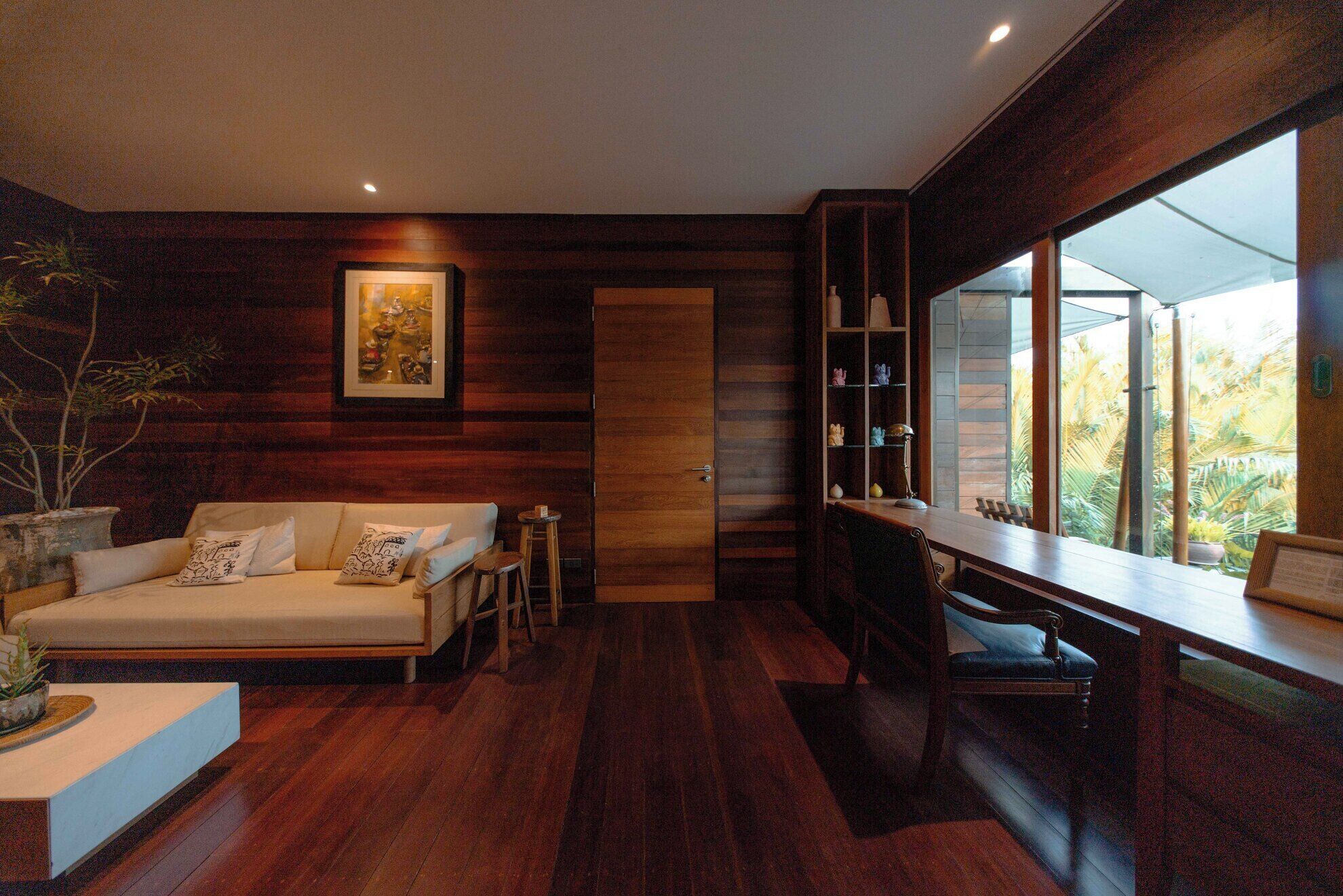 A modern interior with a wooden floor, a sofa, a desk, and a large window offering a view of greenery.