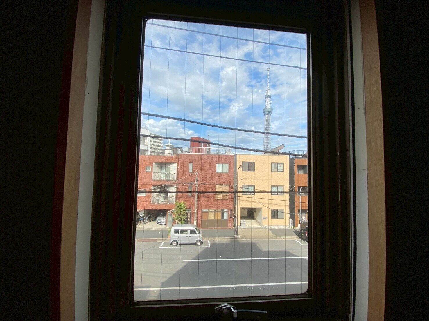 Une fenêtre donnant sur un immeuble à plusieurs étages, une rue et une voiture garée sur le trottoir.