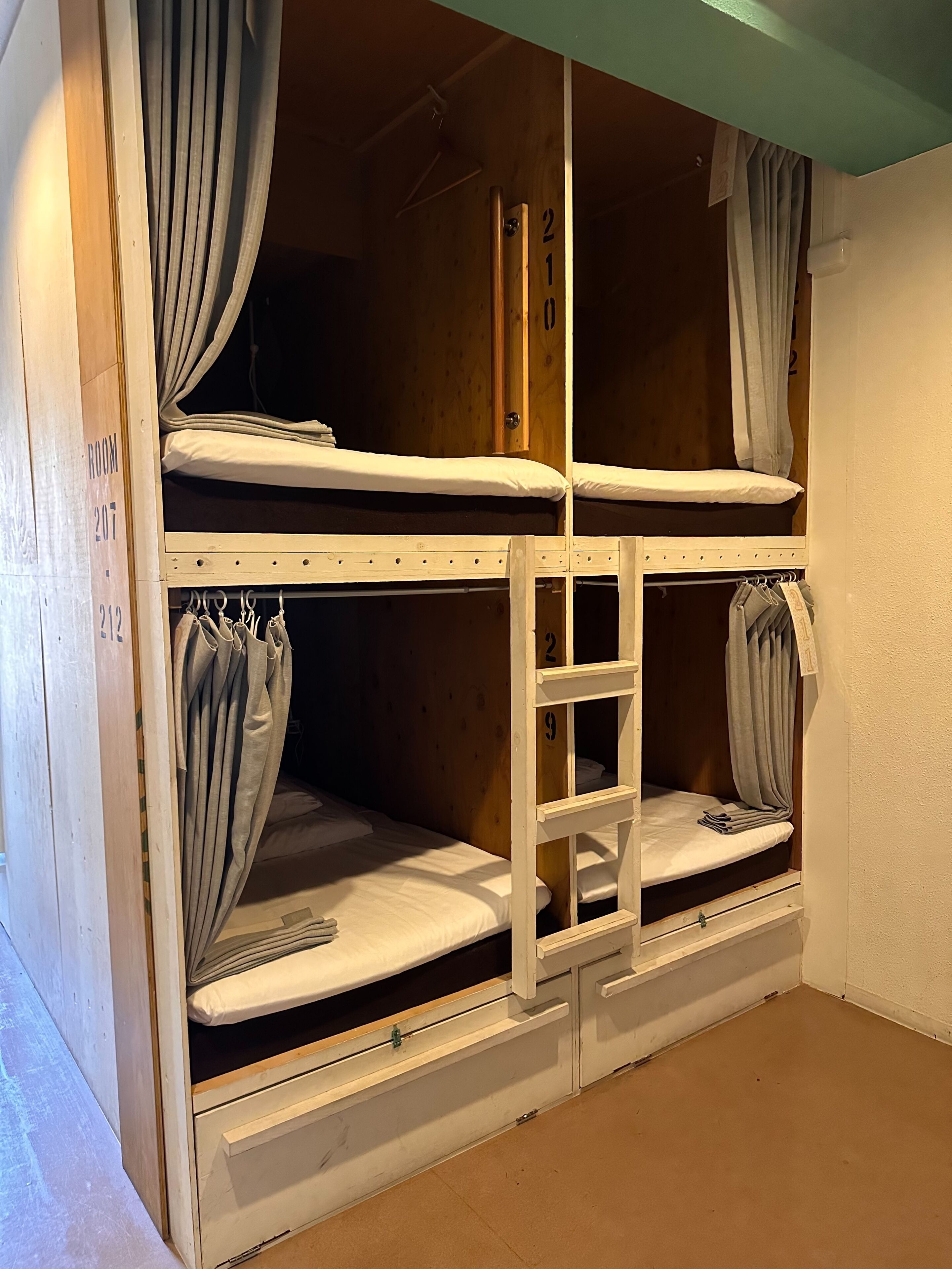 Ein Schlafraum mit einem Etagenbett, drei Ebenen, jeweils mit Vorhang und einer Nummer an der Wand.