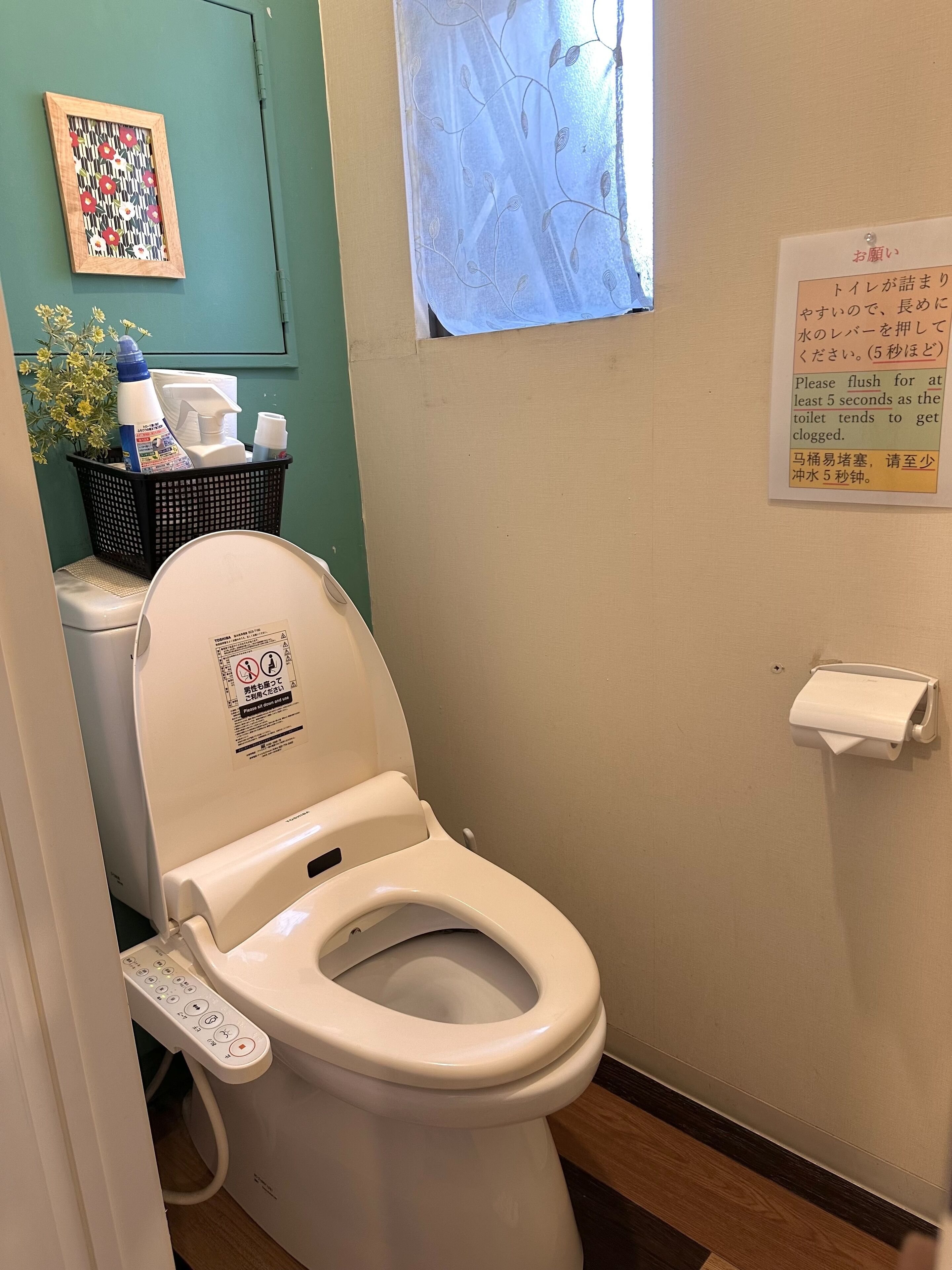 Des toilettes modernes dotées d’un panneau de commande, d’une petite fenêtre agrémentée d’un rideau décoratif et d’une affiche indiquant les instructions en anglais et en japonais.