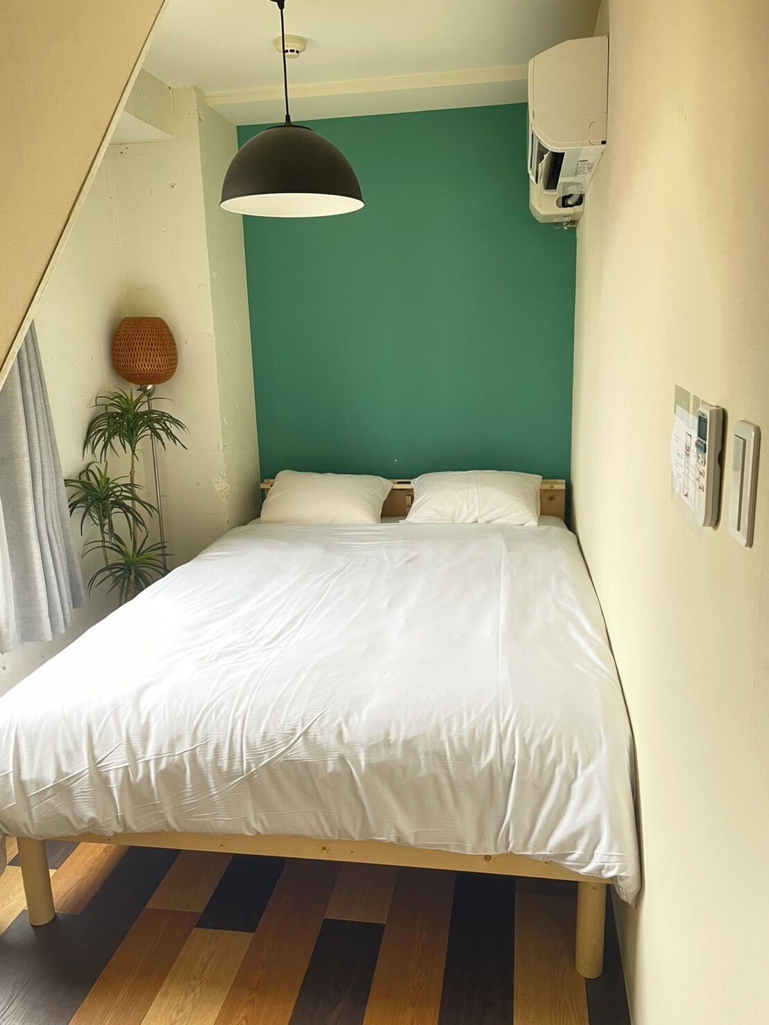 A bedroom with a bed, a hanging lamp, a wall-mounted air conditioner, and a potted plant.