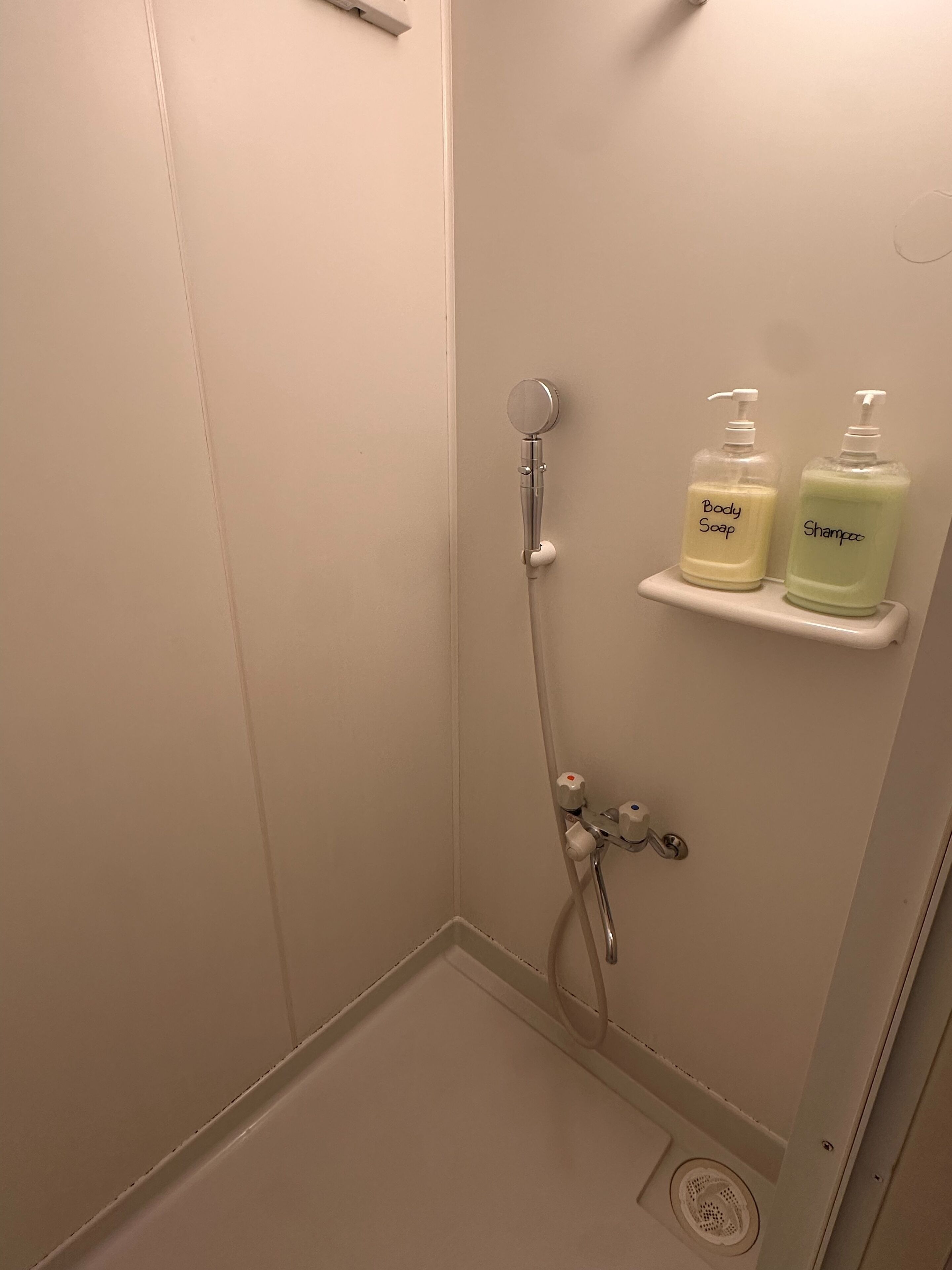A shower with a handheld showerhead, a wall-mounted faucet, and two bottles of body soap and shampoo on a shelf.