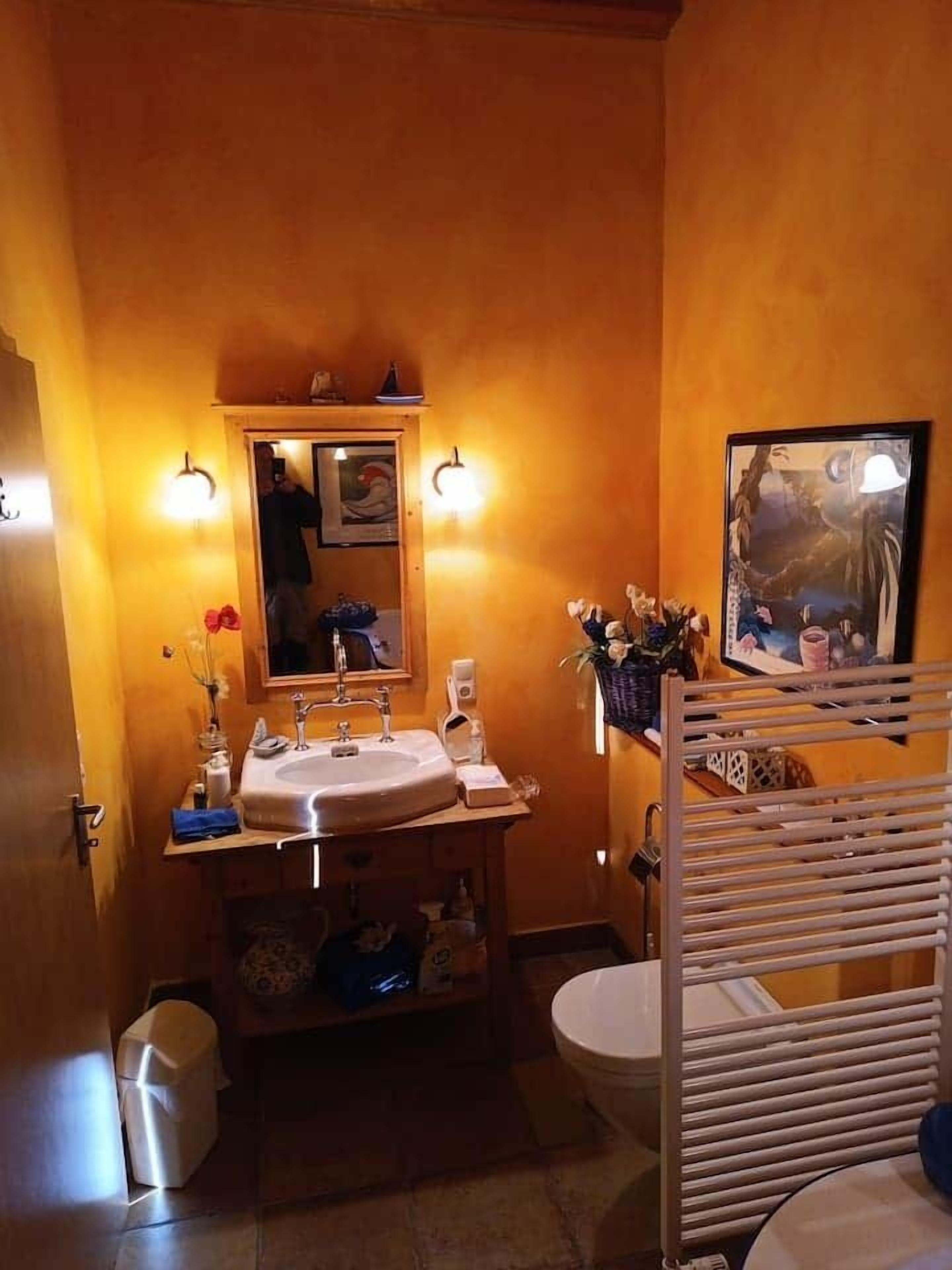 Uma casa de banho com lavatório, espelho e sanita. Há luzes embutidas na parede e um quadro enquadrado.