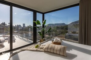 Ein modernes Zimmer mit großen Fenstern, einem Bett, einer Pflanze und Blick auf eine Stadtansicht.
