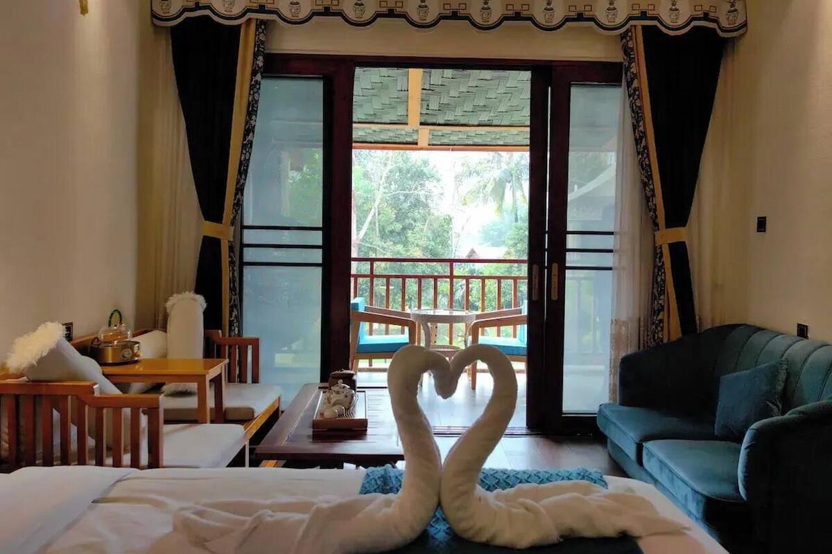 A hotel room with a bed, a sofa, a table with a decorative item, and a heart-shaped towel arrangement on the floor.