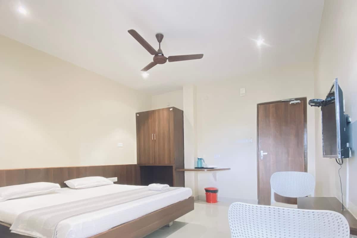 Een moderne hotelkamer met een groot bed, een plafondventilator, een houten garderobe, een klein tafeltje met een waterkoker, een witte stoel en een aan de muur bevestigde televisie.
