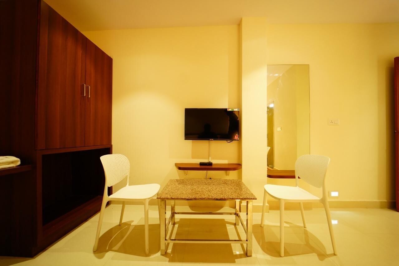 A room with a wooden wardrobe, a TV mounted on the wall, a small table with two white chairs, and a large mirror.