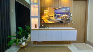 A modern living room with a large TV mounted on a wooden wall, a white entertainment unit below, and a potted plant to the left. - Ella’s Haven @ Shore 3 Residences • 2BR Walk to MOA • Minutes from NAIA (Pasay City)