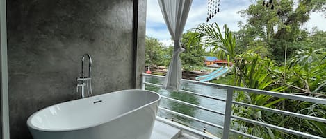 Un baño moderno con una bañera independiente, ducha y vistas a un tobogán acuático y vegetación.