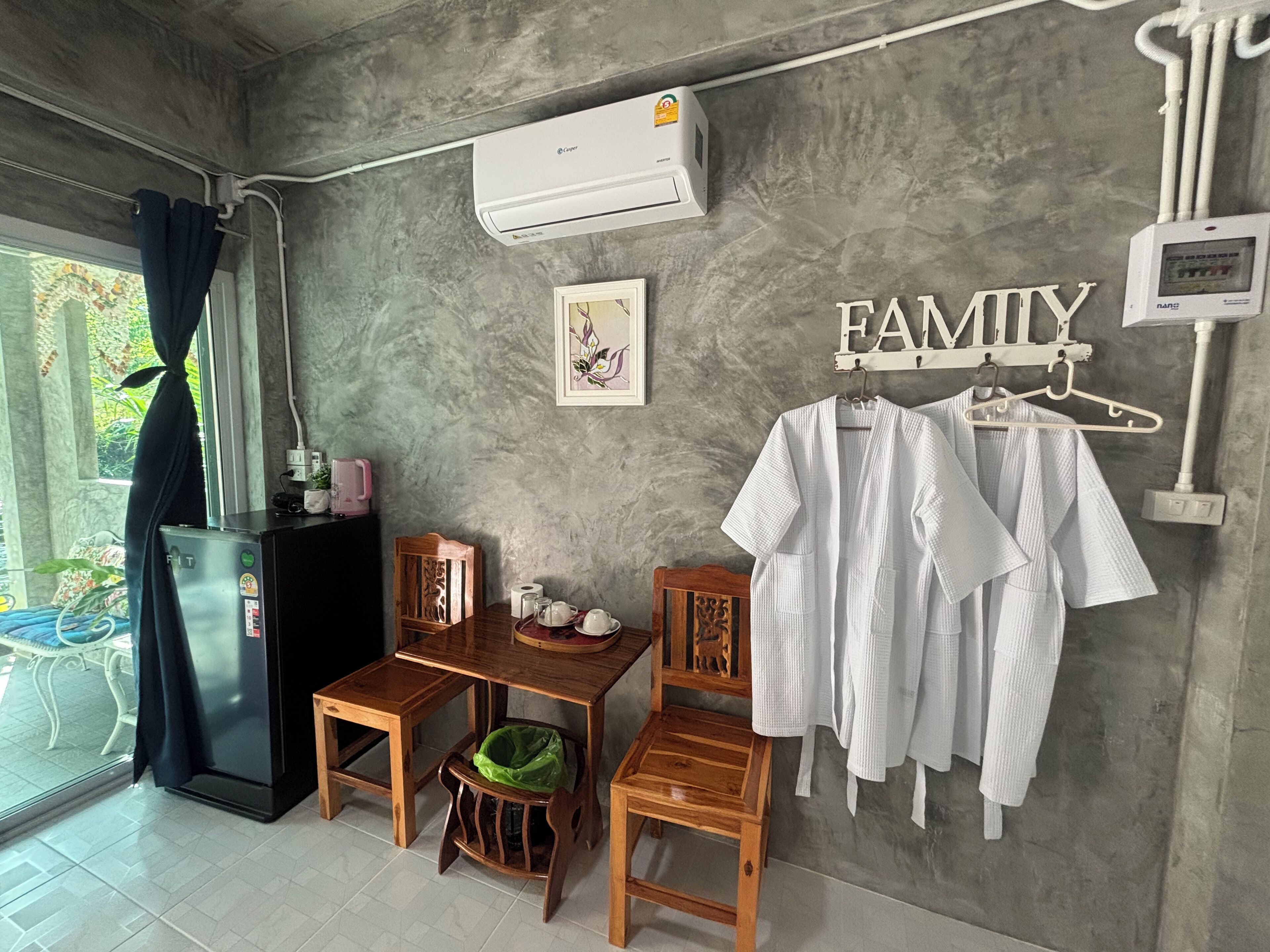 A room with a concrete wall, a small wooden table, a chair, a hanging rack with white shirts, a framed picture, and an air conditioning unit.