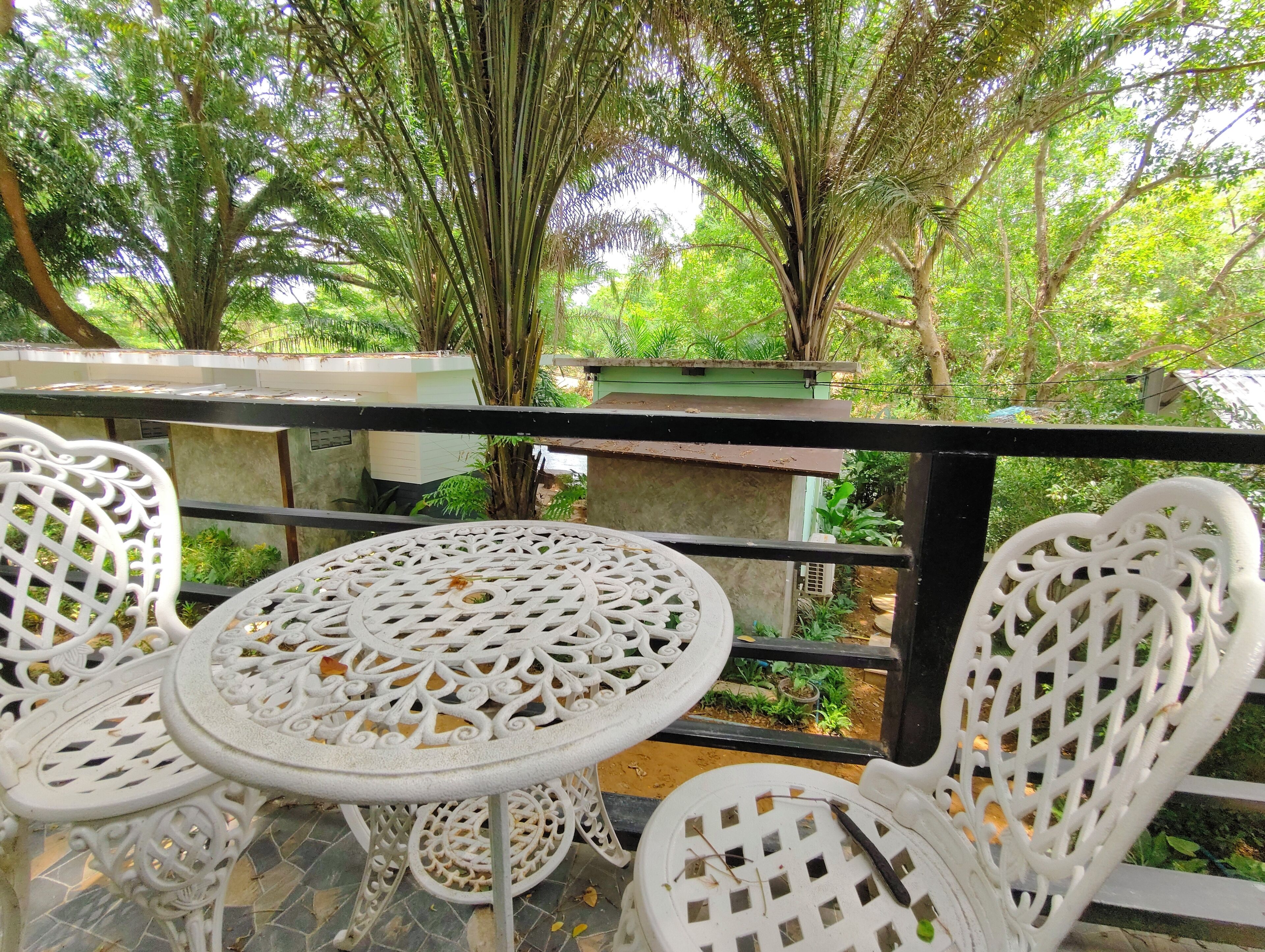 A balcony with white wrought iron chairs and a table, overlooking a lush garden with trees and a green building in the background.