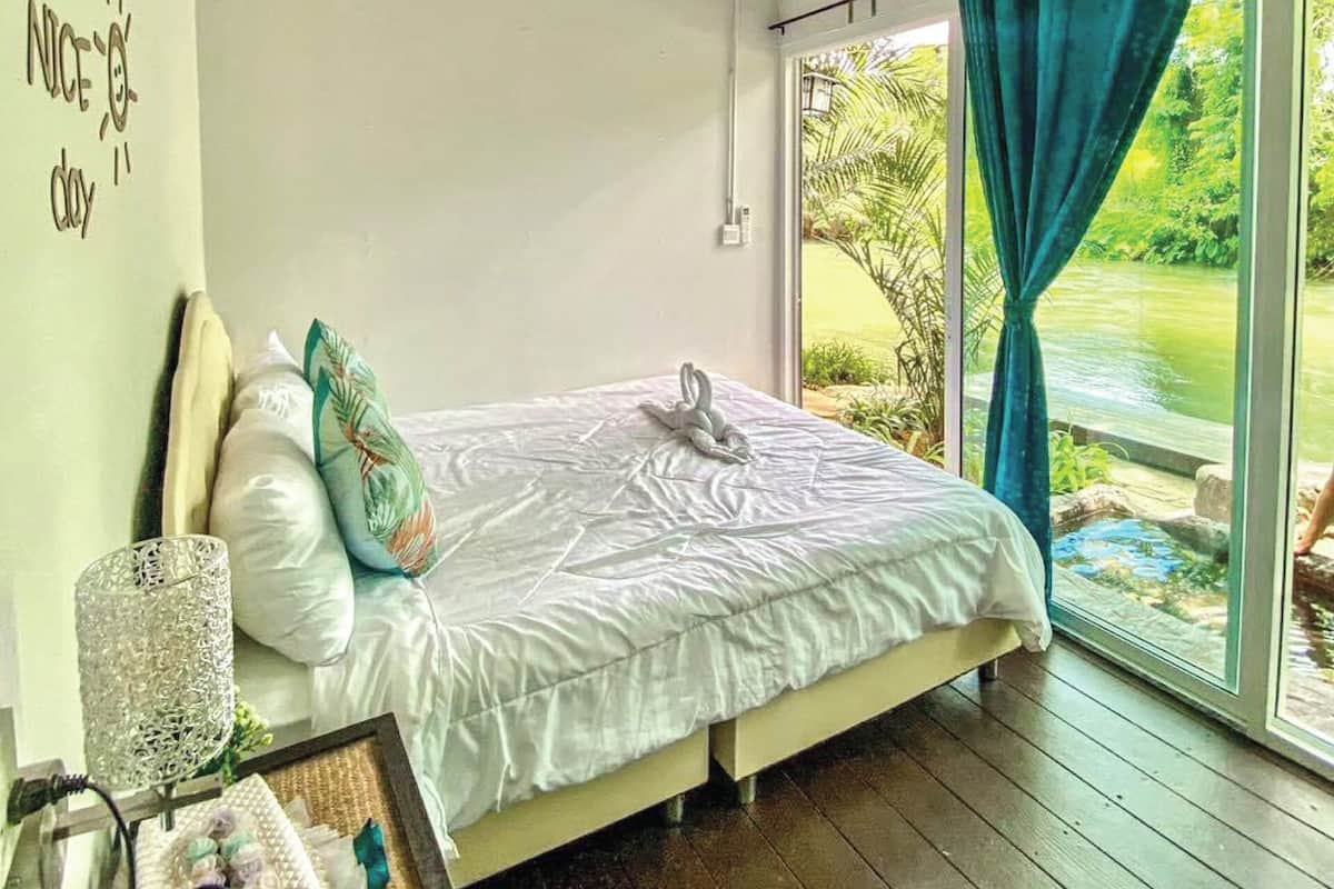 Un dormitorio con cama, almohadas, mesita de noche y ventana con cortinas.