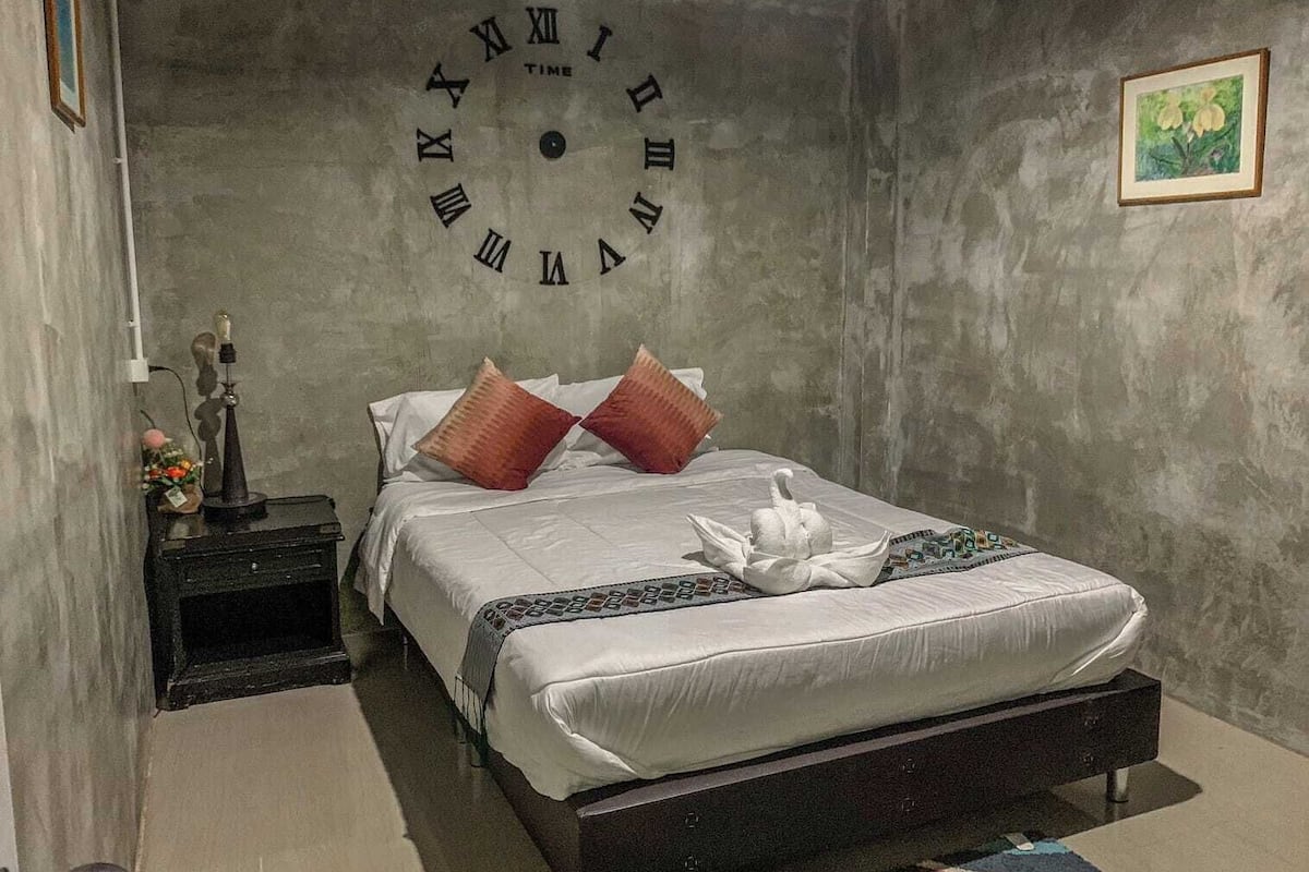 Un dormitorio con un reloj de pared grande, una cama con ropa de cama blanca y almohadas rojas, una mesita de noche con una lámpara, y un cuadro en la pared.