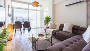 A modern living room with a sofa, a coffee table, and a dining area with chairs and a lamp. - Yellow Canvas 2-BR & Balcony in Larnaca (Larnaca)