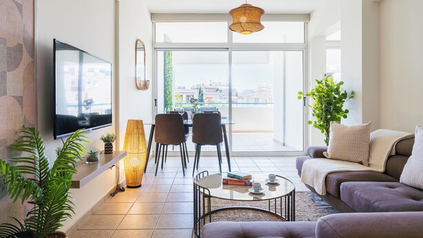 A modern living room with a flat-screen TV, a sofa, a coffee table, and a dining area with chairs and a table. - Yellow Canvas 2-BR & Balcony in Larnaca (Larnaca)
