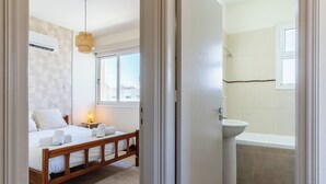 A bedroom with a wooden bed, a bedside table with a lamp, and a view of the city through a window. - Yellow Canvas 2-BR & Balcony in Larnaca (Larnaca)