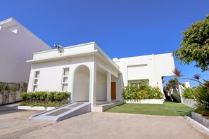 Un edificio moderno con techo plano, entrada cubierta y un pequeño jardín con césped y plantas en macetas.