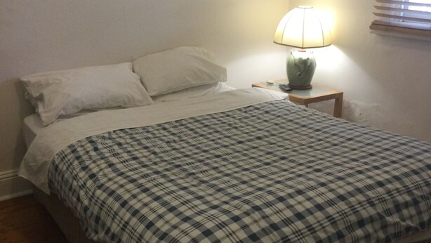 Ein Schlafzimmer mit einem Bett, weißen Kissen, einer karierten Decke, einem Nachttisch mit Lampe und einem Fenster mit Jalousien.