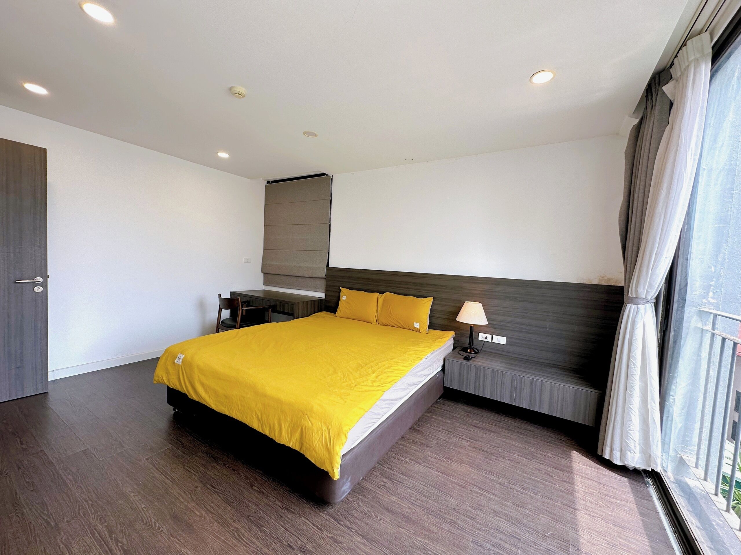 A modern hotel room with a large bed, a desk, a chair, a lamp, and a window with curtains.