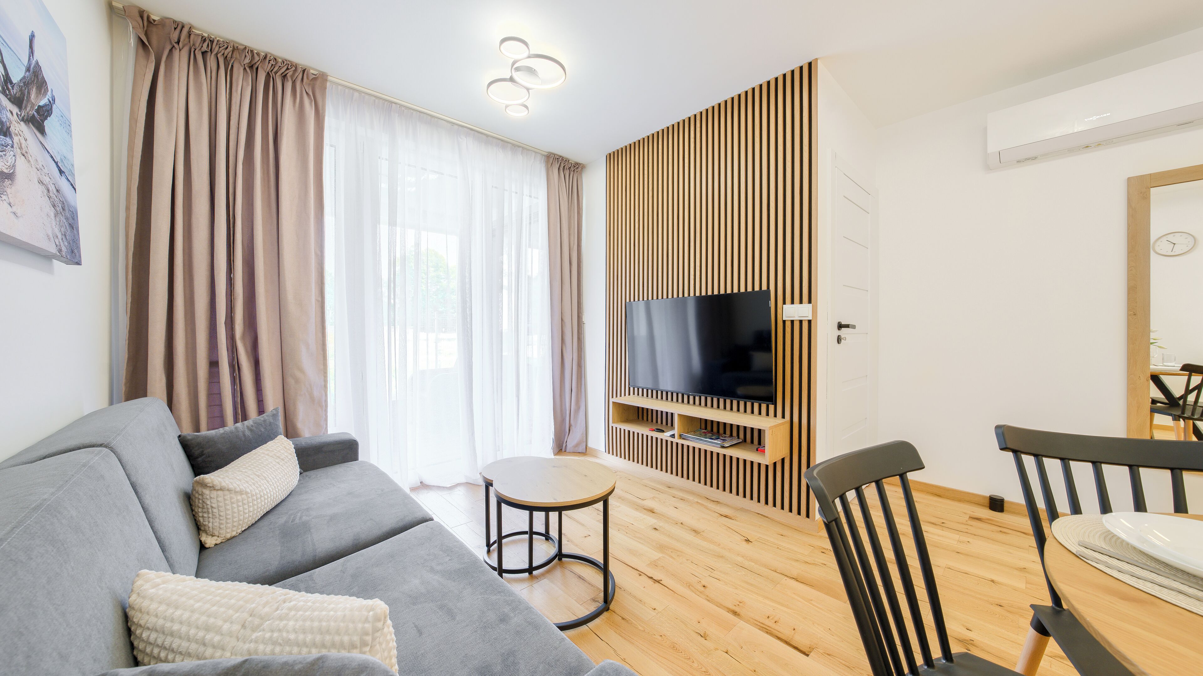 A modern living room with a grey sofa, a small round table, a TV mounted on a wooden panel wall, and a dining area with a wooden table and chairs.