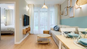 A modern living room with a sofa, a dining table set for two, a TV mounted on the wall, and a large window with curtains. - Apartamenty Sun & Snow Enklawa III (Mielno)