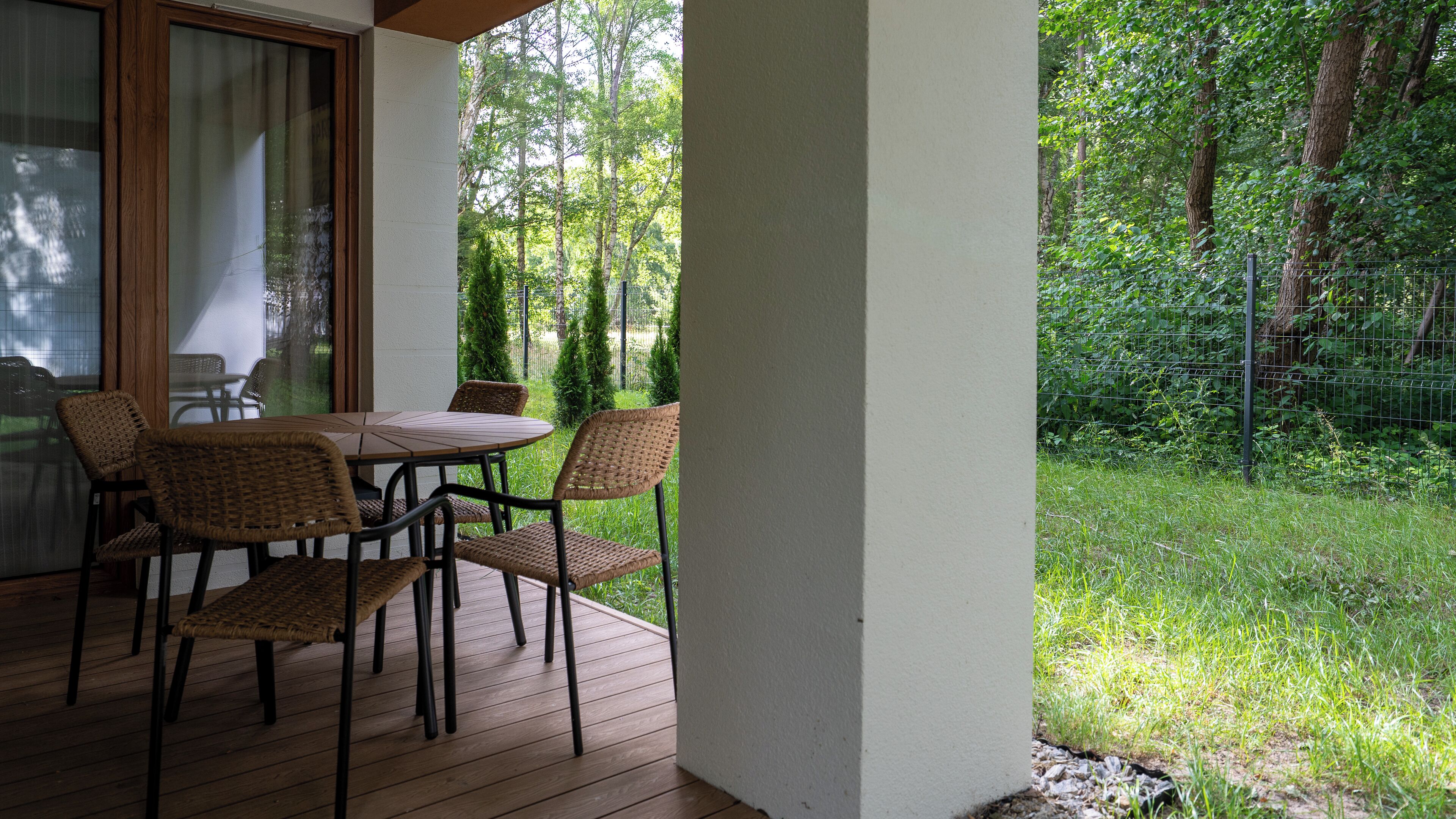 A covered patio with a table and chairs, a view of a green garden, and a wooden deck.