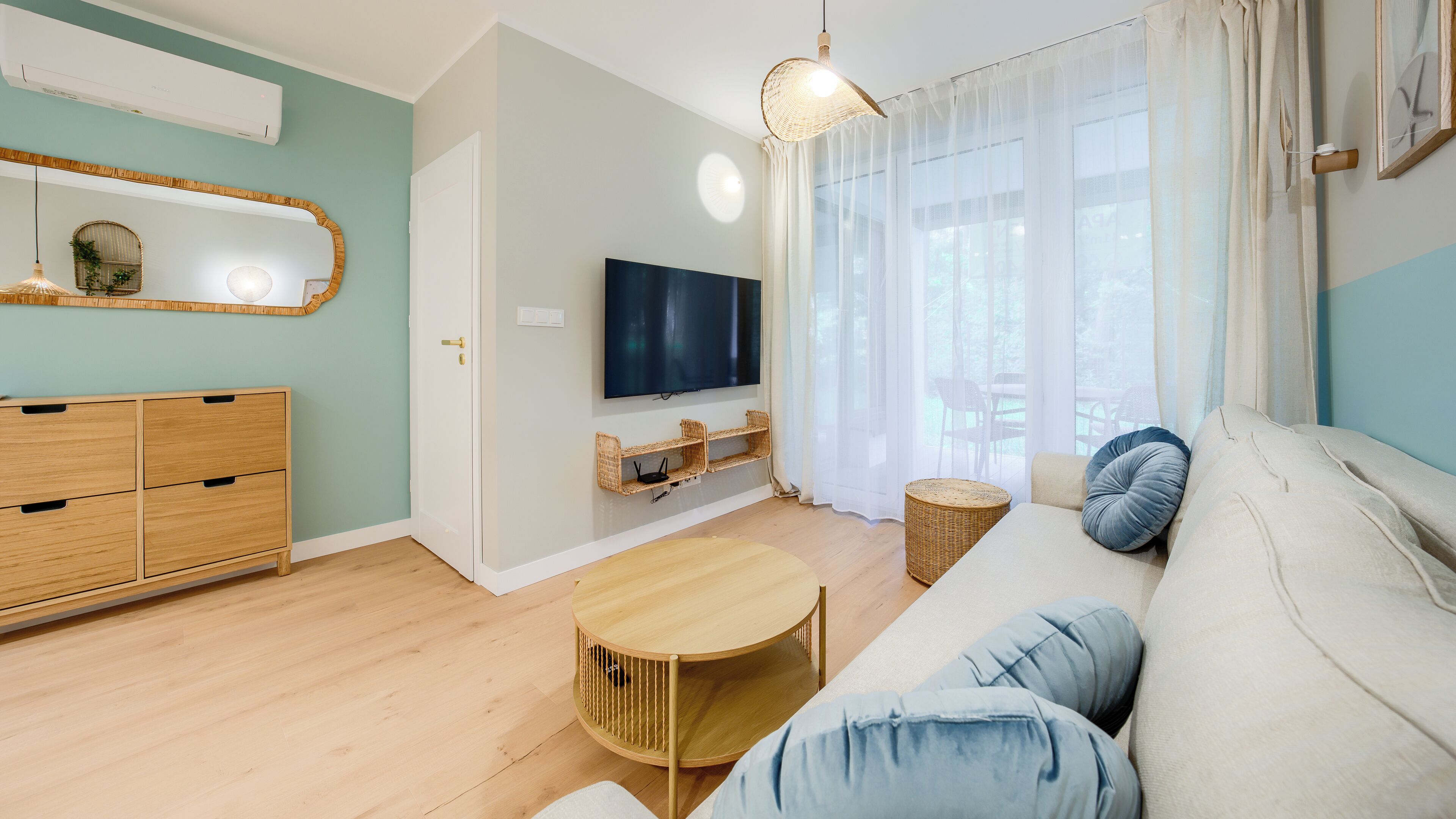 A living room with a TV mounted on the wall, a sofa, a round wooden coffee table, and a wicker side table.