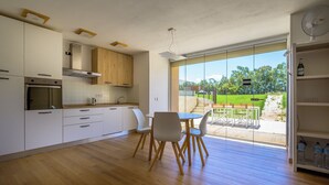 A modern kitchen with white cabinets, a wooden dining table with chairs, and a view of a green garden through large glass doors. - Sunset Villa - Entire Structure (Alghero)