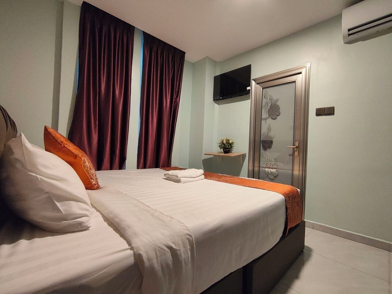 Habitación de hotel con cama, televisor, puerta con panel de vidrio y una mesita con una planta.
