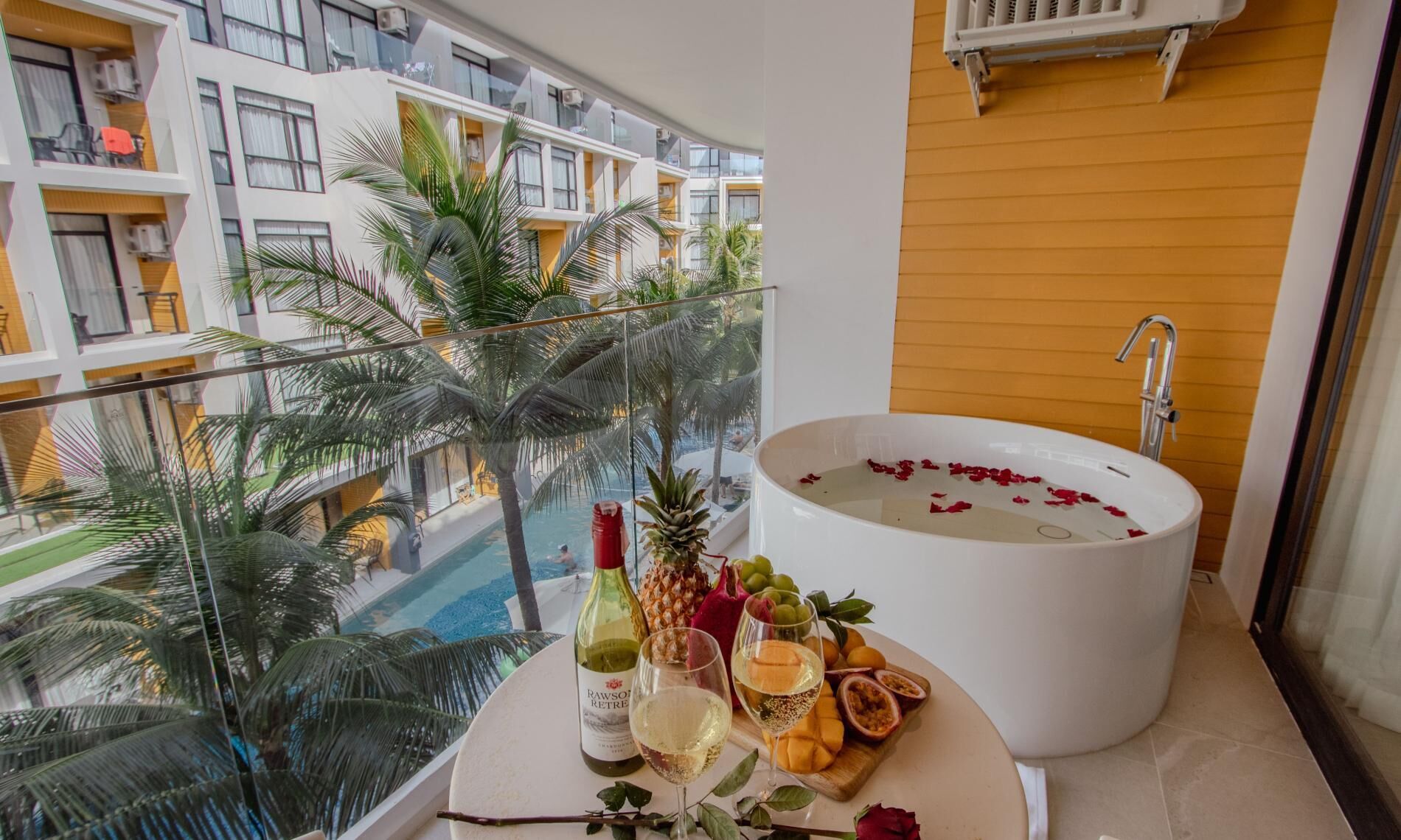 A balcony with a bathtub filled with rose petals, a bottle of wine, and a fruit platter.