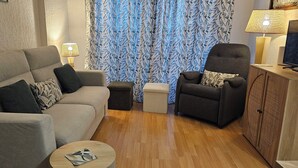 Een woonkamer met een bank, fauteuil, voetenbankje en een kleine tafel. Er staat een lamp en een televisie op een houten kast.