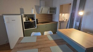 Een praktische keuken met een koelkast, magnetron en spoelbak. Er is een houten tafel met stoelen en een klein aanrecht.