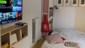 Een slaapkamer met een bed, een televisie op een houten plank, een radiator en een raam met een gestreept gordijn.