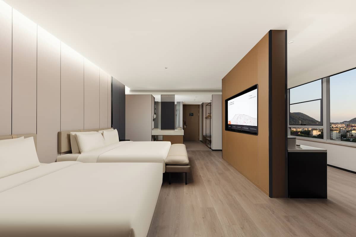 A modern hotel room with two beds, a TV, and a large window offering a city view.
