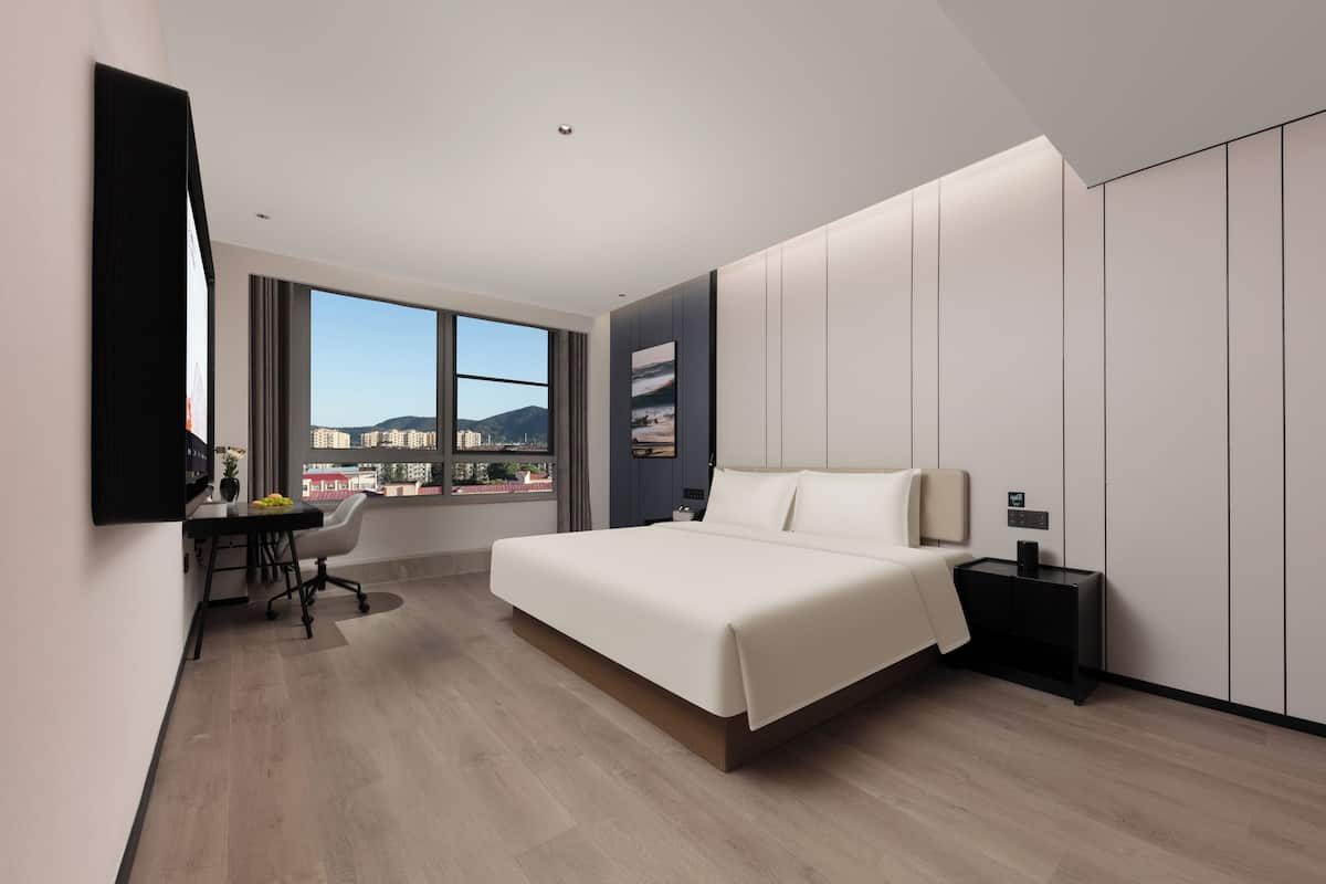 A modern hotel room with a large bed, a desk, and a view of the cityscape.