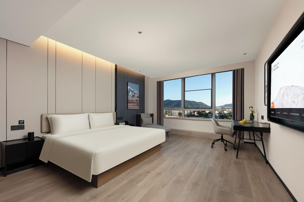 A modern hotel room with a large bed, a desk, and a view of the cityscape through large windows.
