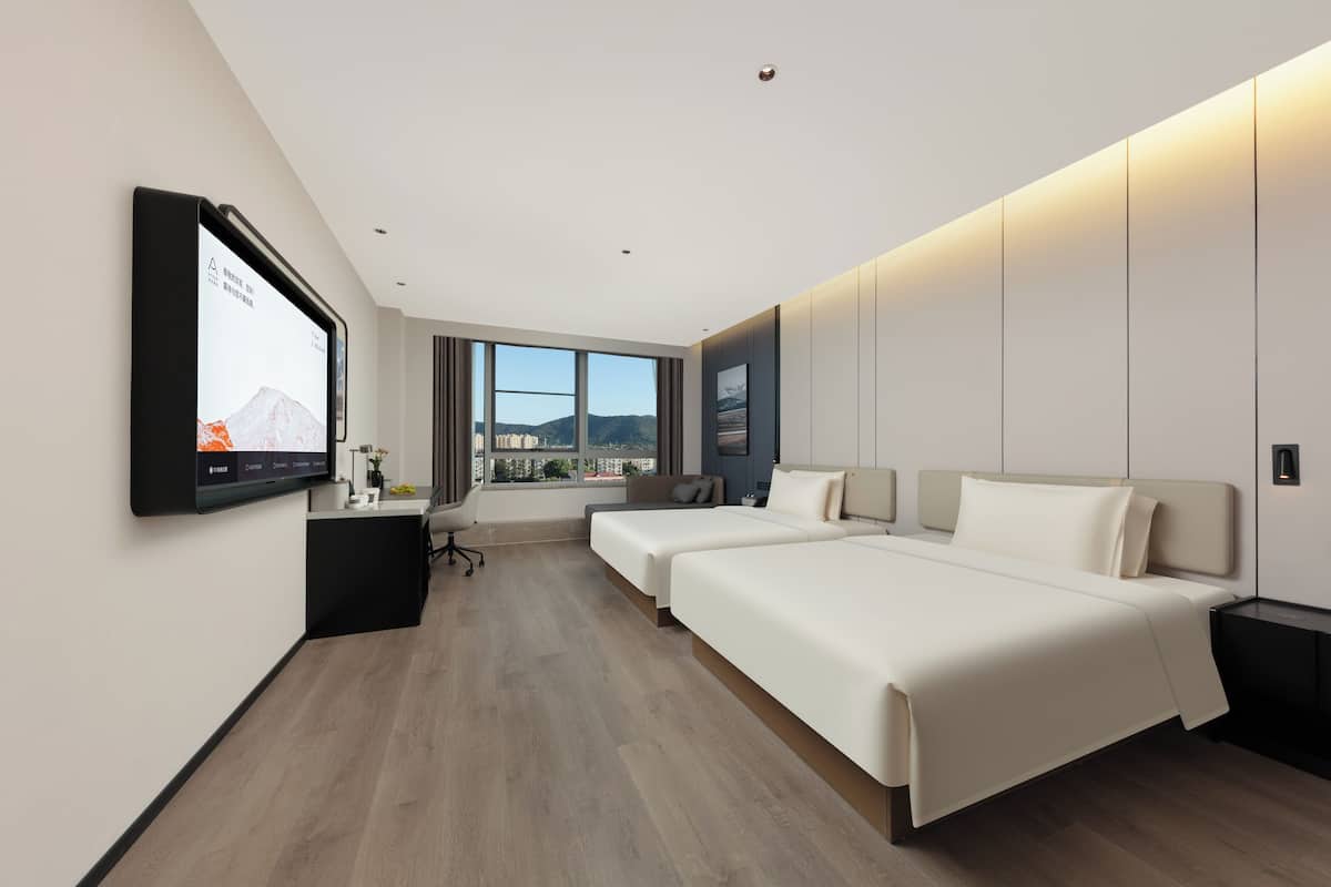 A modern hotel room with two beds, a desk, a chair, and a large window offering a city view.