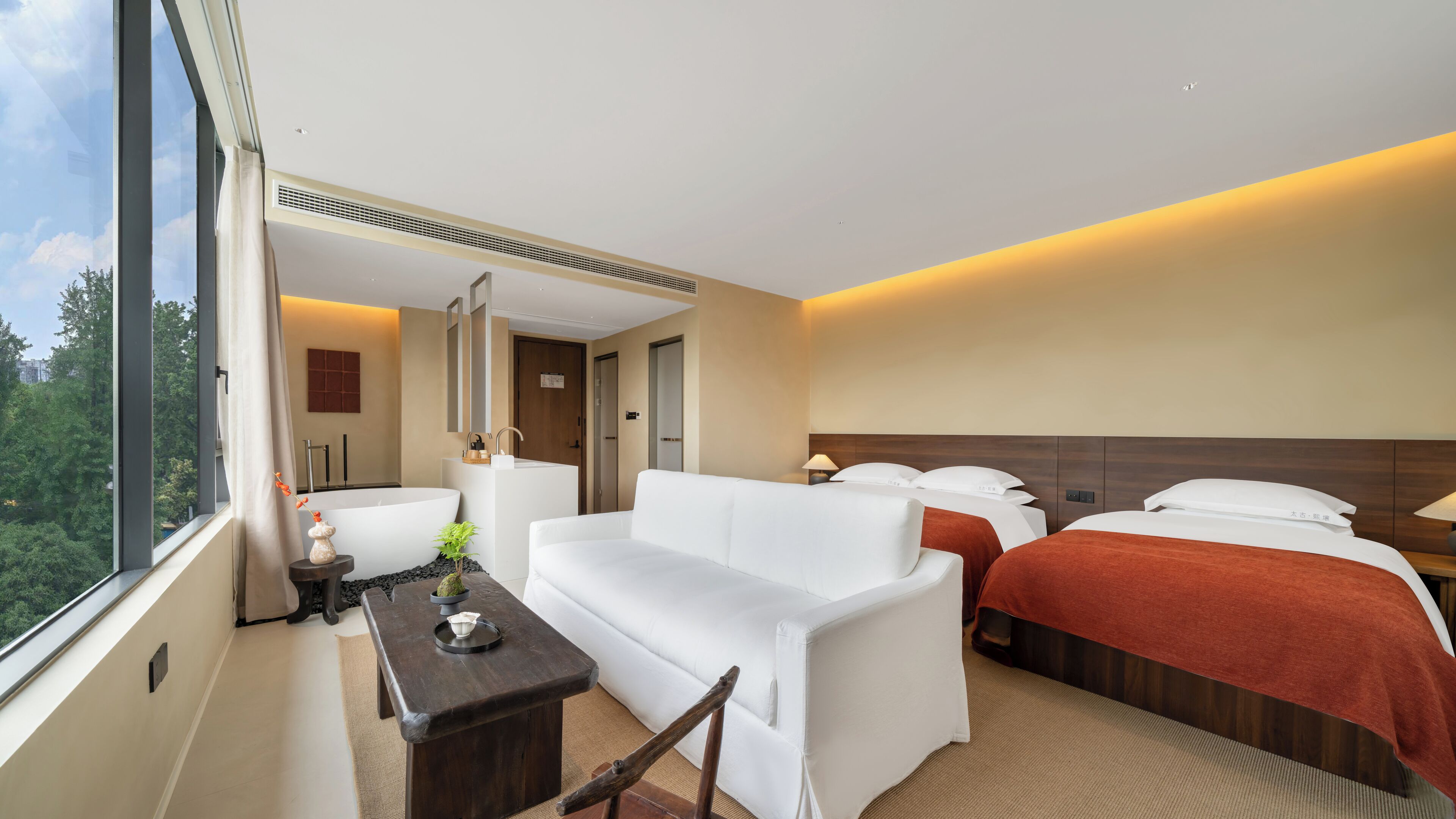 Habitación de hotel con un ventanal grande, un sofá blanco, dos camas con ropa de cama roja, una mesita con una planta y un espejo sobre el sofá.