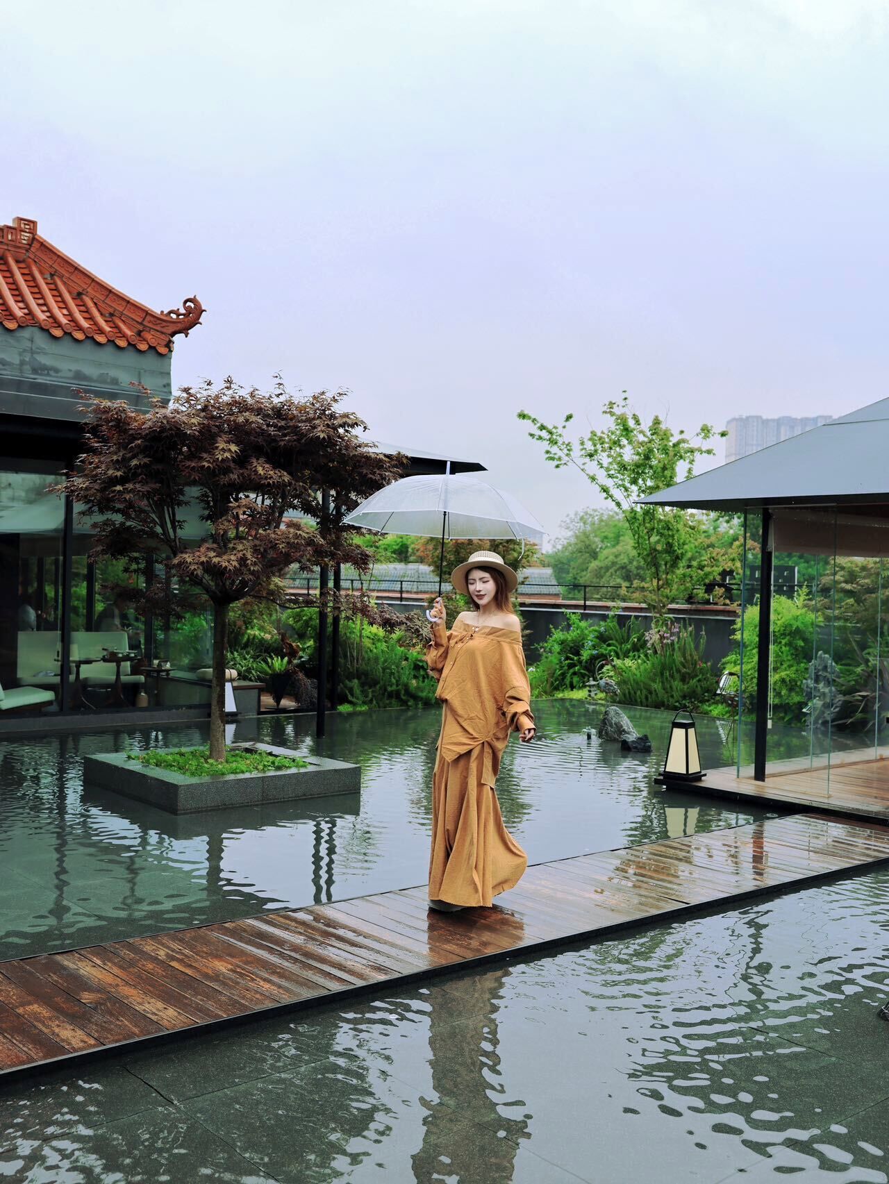 A person standing on a wooden deck with a transparent umbrella, surrounded by water and greenery.