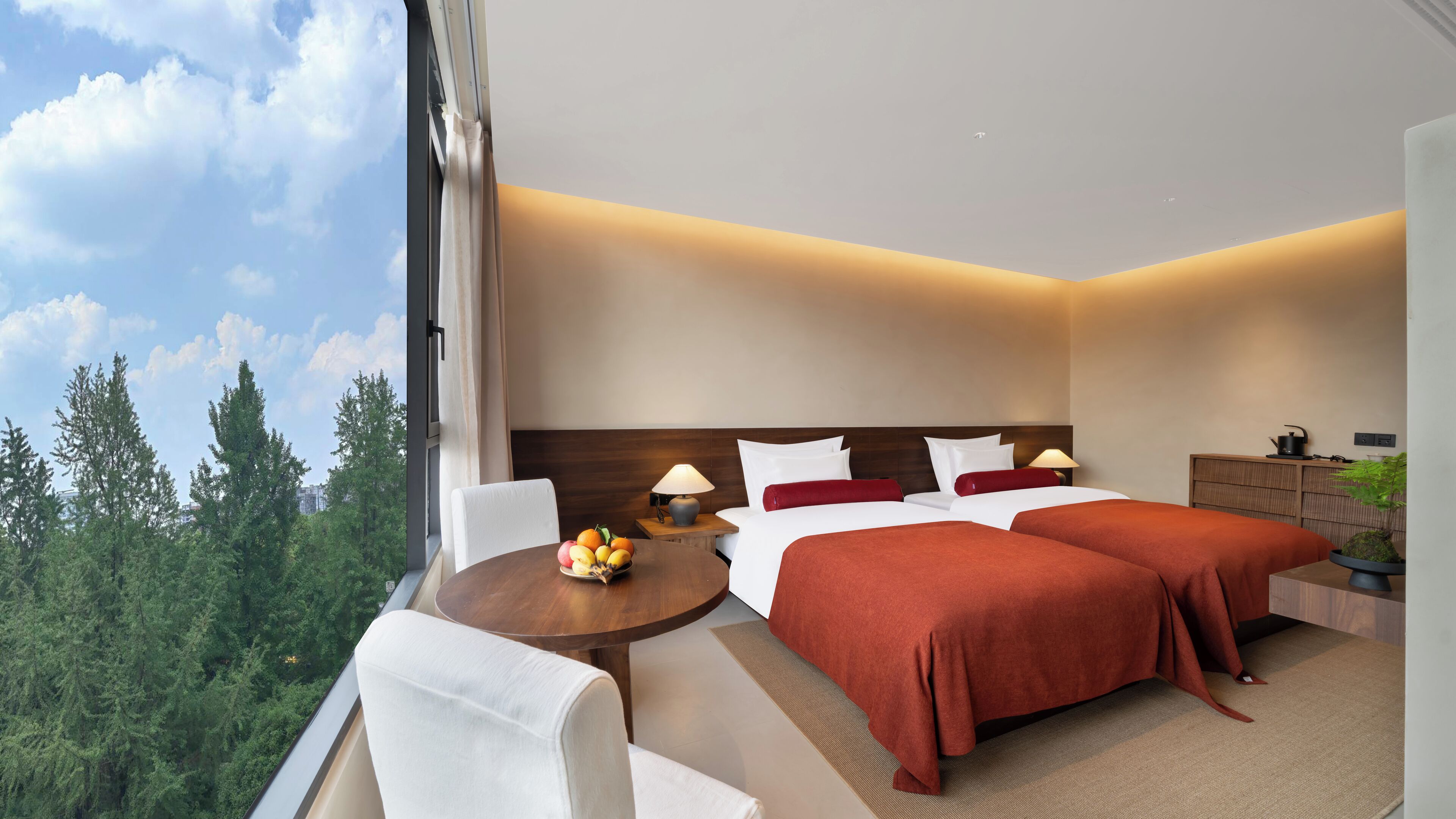 Habitación de hotel con una cama grande, dos mesitas de noche, una pequeña mesa redonda con fruta y vistas a árboles.