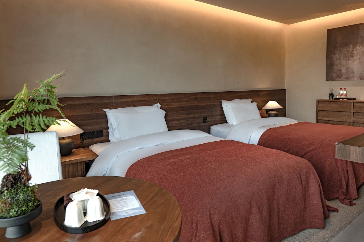 Camera d'albergo con due letti, una testiera in legno, un comodino con una lampada, un tavolino con una pianta e un portatovaglioli, e una mensola a parete con una bottiglia e una scatola.