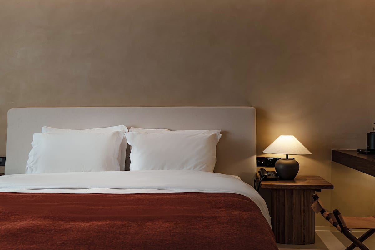 Una camera d'albergo con un letto grande, un comodino in legno, una lampada e una sedia.