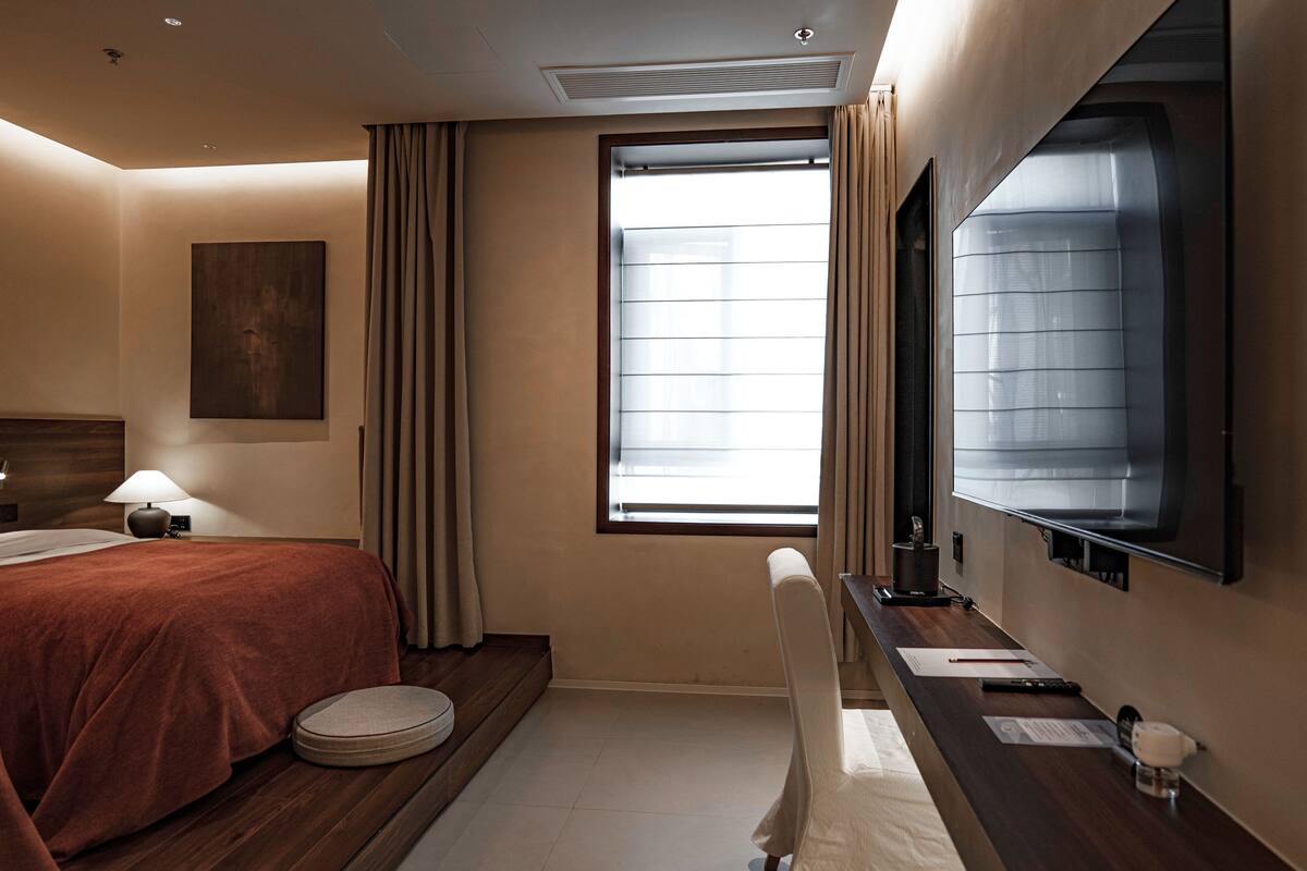 Una camera d'albergo con un letto, una scrivania con una sedia, un'ampia finestra con tende, una TV a muro e un'opera d'arte appesa.