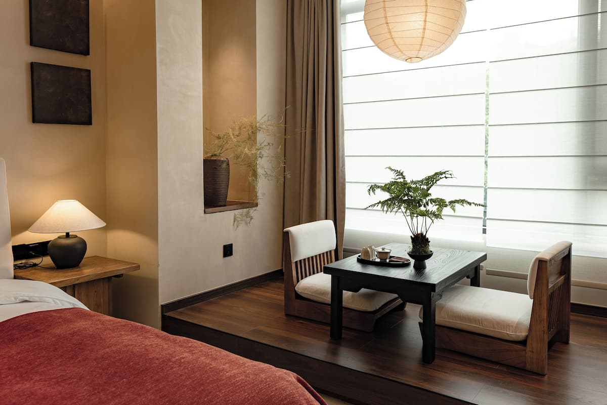 Una camera d'albergo con pavimento in legno, un letto con coperta rossa, un tavolo con una pianta, una sedia e un lampadario.