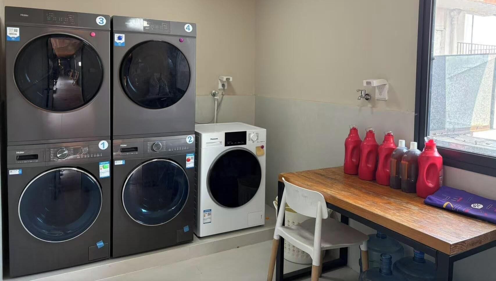 Une buanderie équipée de quatre machines à laver et de quatre sèche-linge, une table avec des produits de nettoyage et une fenêtre donnant sur l’extérieur.